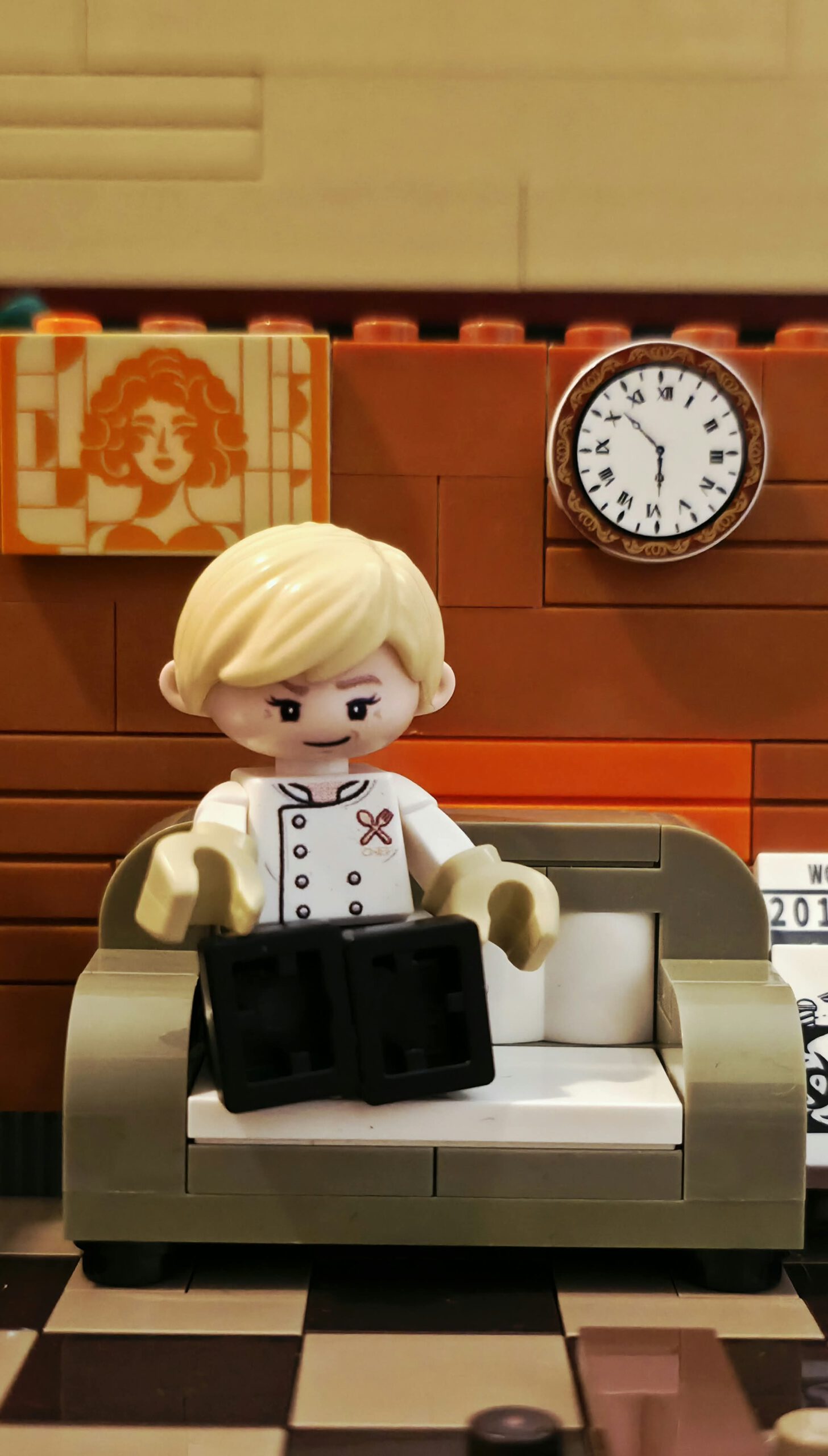 Blonde IAMBRICK Kochfigur auf einem Sofa vor dekorierter Wand und runder Uhr